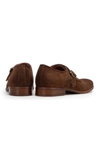 Zapatos de ante marrón con una textura suave, puntera redonda y detalle de hebilla decorativa; acabados con un tacón de madera medio y una silueta limpia.