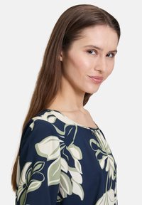 Granatowa bluzka w kwiaty z zielonymi i kremowymi wzorami liści, okrągły dekolt, miękka tkanina i luźny krój. Posiada delikatne marszczenie przy dekolcie.