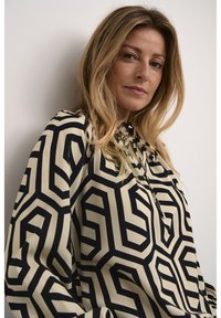 Blusa astratta a motivi color crema e nero. Caratterizzata da forme geometriche, maniche lunghe e un collo arricciato con dettaglio a fiocco.