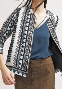 Zwart-witte gestreepte blazer met geometrische patronen, gecombineerd met een blauwe top en een bruine rok, met een rits aan de voorkant en driekwart mouwen.