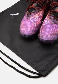 Des crampons de soccer violets et noirs avec un motif en rayures, placés sur un sac à cordon noir arborant un logo blanc.