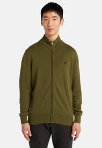 Olivgrön zip-up tröja i stickad kvalitet, med hög krage, ribbade ärmslut och nederkant, samt en liten svart logotyp på bröstet.