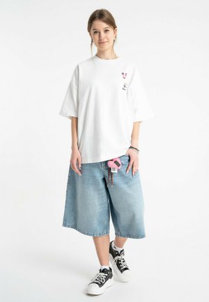 Młoda kobieta stojąca, ubrana w oversize'ową białą koszulkę, długie szorty jeansowe oraz czarno-białe trampki na białym tle.