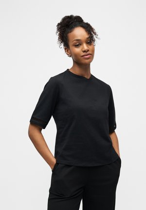 Ung kvinne med krøllete hår samlet opp, iført svart kortermet skjorte og svarte bukser, poserer med hendene i lommene foran en hvit bakgrunn.