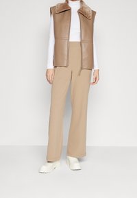 Brązowa bezrękawnik skórzany z futrzanym kołnierzem, w połączeniu z beżowymi szerokimi spodniami i masywnymi białymi botkami za kostkę, na jednolitym tle.