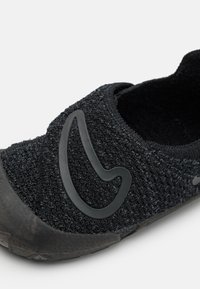 Svart slip-on-sko med övre del i texturerad tyg, gummisula och en framträdande svart logotypdetalj på sidan.