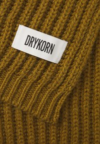 Tejido de punto acanalado color mostaza con una etiqueta blanca visible que dice "DRYKORN" en letras negras en negrita. Textura suave y costuras prominentes.