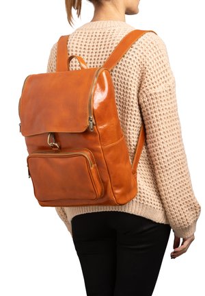 Persoon draagt een gebreide beige trui en een zwarte broek, met een grote lichtbruine leren rugzak met ritsvakken en een sluiting met gespen.