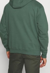 Sudadera con capucha de color verde oscuro con una textura suave, que cuenta con capucha, puños y dobladillo acanalados. Se combina con pantalones verde oscuro, mostrando un diseño sencillo.