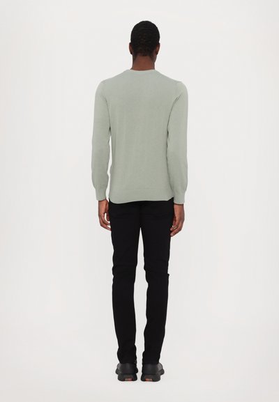 Sweater verde claro de mangas longas, com punhos e bainha canelados, combinado com calças pretas e sapatos pretos, visto de costas.