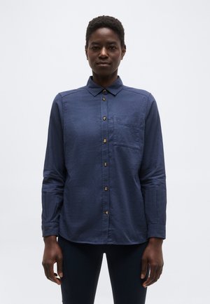 Άτομο με κοντά μαλλιά που φοράει σκούρο μπλε μακρυμάνικο πουκάμισο με κουμπιά και σκουρόχρωμο παντελόνι, στέκεται μπροστά σε απλό φωτεινό φόντο.