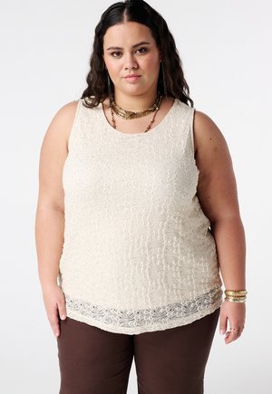 Vrouw met lang donker haar, gekleed in een mouwloze crèmekleurige kanten top, bruine broek, gouden ketting, oorbellen en armbanden, staand tegen een effen achtergrond.