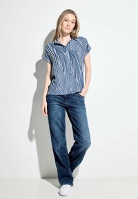 Camicia a maniche corte con bottoni, con motivo a righe blu e bianche, abbinata a jeans scuri larghi e sneakers bianche.
