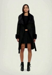 Femme portant un manteau noir avec un col en fourrure, une jupe noire, des bottines, et un tatouage "1999" visible sur sa cuisse droite, se tenant devant un fond uni.