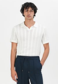 Gestreiftes Strickpoloshirt in Weiß mit strukturiertem Muster und kurzen Ärmeln. Kombiniert mit dunkelblauen Hosen mit elastischem Bund und Taschen.