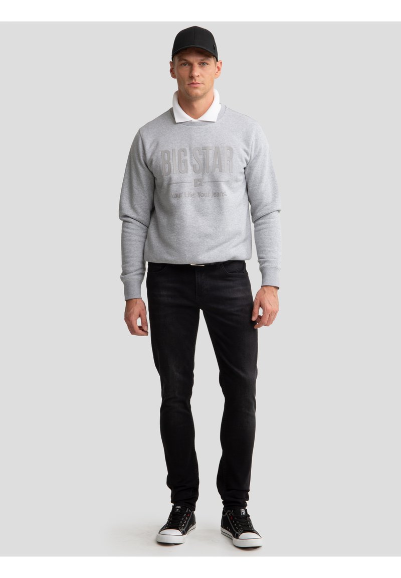 Mand iført sort kasket, grå sweatshirt med teksten "BIG STAR", sorte jeans og sorte sneakers, stående foran en lys grå baggrund.