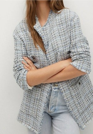 Femme portant un blazer en tweed bleu clair avec des bords effilochés et des manches retroussées, associé à un jean délavé clair, bras croisés.