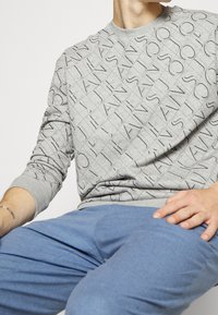 Šedá mikina s prošívaným vzorem, která obsahuje opakující se textové vzory v tmavší šedé. Nosit s světle modrými kalhotami.