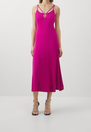 Frau trägt ein ärmelloses magentafarbenes Midikleid mit dünnen doppelten Trägern und geschnürten High-Heel-Sandalen vor einfachem Hintergrund.