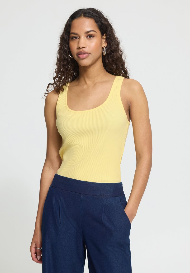 Femme aux cheveux bouclés portant un haut sans manches jaune et un pantalon bleu marine, debout avec une main dans la poche sur fond blanc.