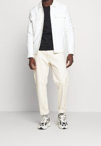 Chaqueta blanca abotonada sobre una camisa negra, combinada con pantalones crema claro y zapatillas multicolores. Las telas parecen suaves con líneas estructuradas.