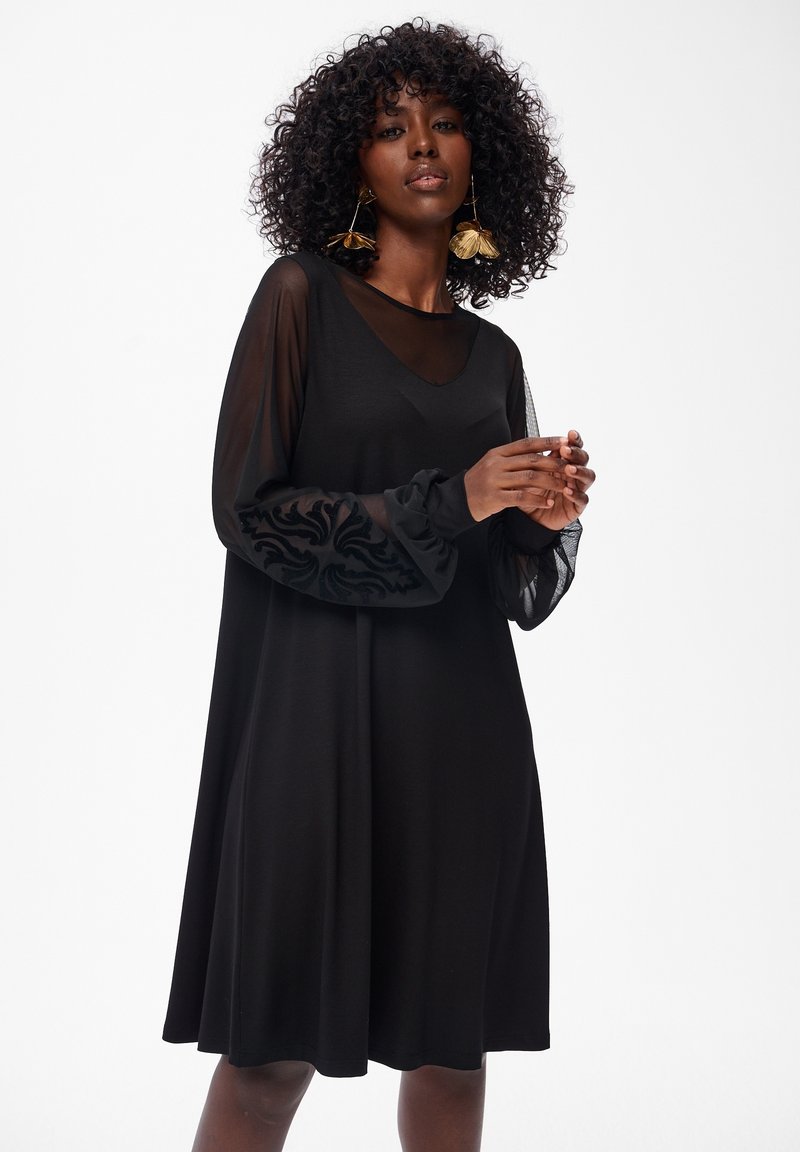 Frau mit lockigem Haar, die ein schwarzes Kleid mit durchsichtigen, bestickten Ärmeln und großen goldenen Blumenohrringen trägt, posiert vor einem weißen Hintergrund.