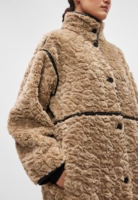 Bežīgs plīša mētelis ar augstu apkakli, melni pogu aizdari un kontrastējošu apmali gar šuvēm. Tekstūras virsma ar mīkstu izskatu.