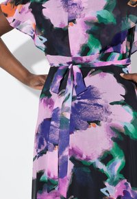 Robe à motif floral avec une palette de couleurs mauve, verte et noire, en tissu doux, avec des manches fluides et un détail de taille nouée.