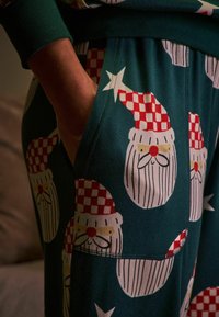 Zelené pyžamové kalhoty vyrobené z látky s opakujícím se vzorem obličejů Santa Klauze, kteří mají červené kostkované čepice, bílé vousy a žluté akcenty.