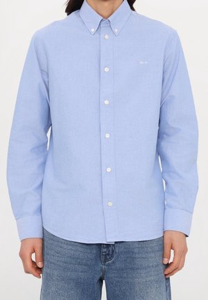 Homme portant une chemise bleu clair à manches longues avec un petit texte blanc "NN.07" sur la poitrine, associée à un jean bleu.
