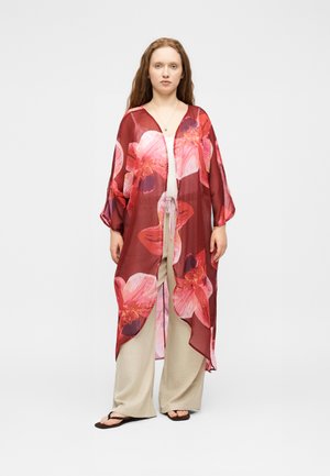Nő hosszú, áttetsző piros kimonót visel nagy rózsaszín virágmintákkal, bézs színű bőszárú nadrággal, fehér toppal és fekete szandállal, fehér háttér előtt áll.