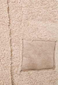 Krämfärgad fluffig tyg med en strukturerad, lockig yta; har en slät, mockaliknande ficka.