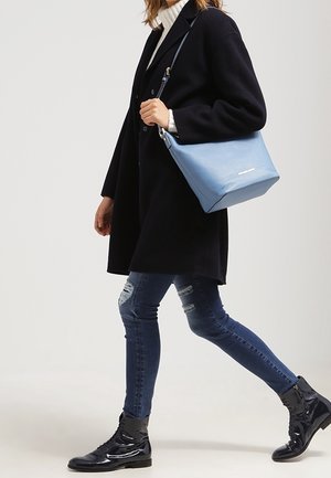 Borsa a tracolla in pelle blu con finitura testurizzata, forma rettangolare e logo argento, abbinata a un lungo cappotto nero e jeans.