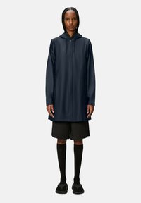 Giacca da pioggia navy con zip frontale, cappuccio regolabile e due tasche laterali, abbinata a pantaloni corti neri e calze fino al ginocchio.