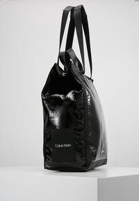Czarna, błyszcząca torba tote z podwójnymi czarnymi uchwytami, z prostokątną naszywką z logo "Calvin Klein". Gładka tekstura o minimalistycznym wzornictwie.
