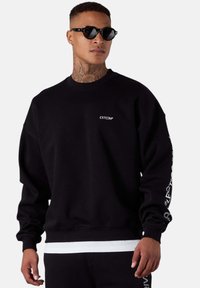 Sweatshirt noir avec une coupe décontractée, des poignets côtelés et un col rond. Présente un branding blanc sur la poitrine et le long de la manche. Porté avec des lunettes de soleil.