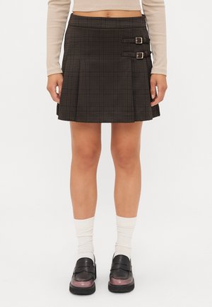Kvinde iført et mørkt ternet plisseret skørt med to spænder i siden, hvide sokker, sorte loafers og en langærmet beige cropped top.