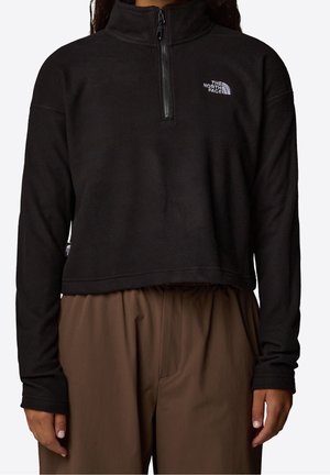 Sort fleece-trøje med lange ærmer, kvart lynlås og logo på venstre bryst, kombineret med løse brune bukser på en stående person.
