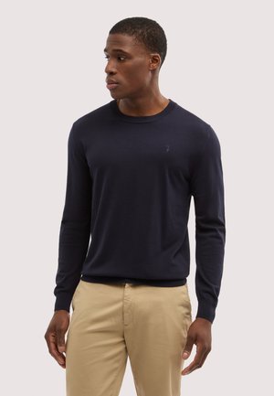 Mann trägt einen marineblauen Langarmpullover und beige Hosen, schaut nach links vor einem schlichten, hellen Hintergrund.