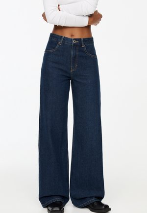 Persona que lleva vaqueros anchos de color azul oscuro y zapatos negros, con los brazos cruzados sobre un top blanco corto de manga larga.