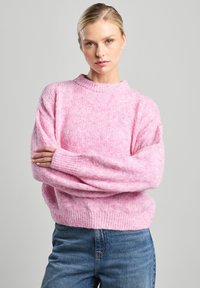 Pink, textured knit sweater with a round neckline and ribbed cuffs. Worn with blue jeans, featuring loose sleeves and a relaxed fit.