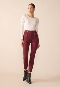 Pantalons rouges taille haute avec des accents de boutons argentés, silhouette ajustée et détails sur les panneaux latéraux, associés à un haut ajusté à manches longues blanc.