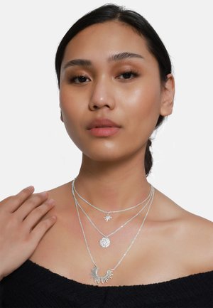 Colliers superposés en argent avec différents pendentifs : une étoile, un cercle texturé et un design en rayons de soleil. Portés sur une peau nue.