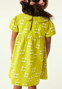 Bambina con i capelli ricci legati in fiocchi rosa, che indossa un vestito verde a maniche corte con fantasia di fiori bianchi, rivolta di spalle rispetto alla fotocamera.