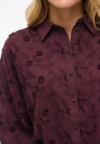 Chemise boutonnée bordeaux avec broderie florale, col classique, et tissu texturé avec des motifs en relief de fleurs et de feuilles.