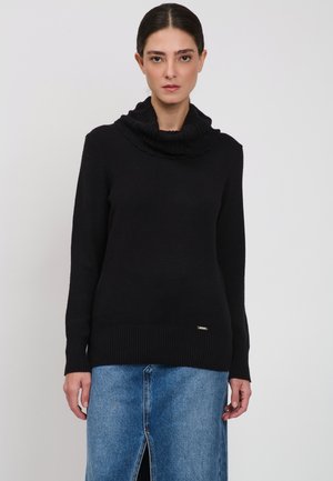 Kvinde iført en sort strikket sweater med flæset rullekrave og en blå denimsnørret nederdel med slidse foran, stående mod en hvid baggrund.