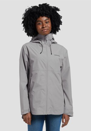 Vrouw met krullend haar die een lichtgrijze regenjas met capuchon draagt, dichtgeritst, met handen ontspannen langs haar zij, tegen een effen achtergrond.