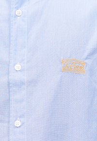 Φωτεινό γαλάζιο και λευκό ριγέ πουκάμισο με λευκά κουμπιά και κίτρινο κεντημένο λογότυπο που μοιάζει με κτίριο με τη λέξη "Agless".