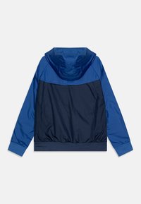 Modrá větruvzdorná bunda s kapucí, která má tmavě modrou tělesnou část a světle modré rukávy. Vyrobena z hladkého, lehkého materiálu s elastickými manžetami.