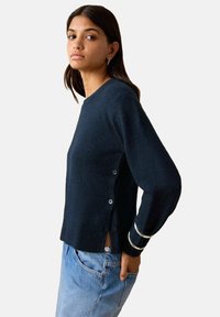 Femeie îmbrăcată într-un pulover bleumarin închis cu detalii cu nasturi pe lateral și blugi albastre deschiși, pozând cu o mână în buzunar, pe un fundal alb.